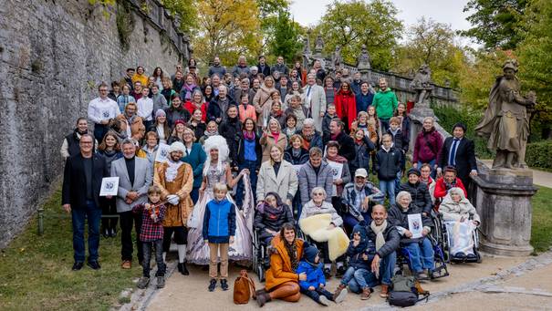 Auf einer Treppe in einem Park oder Garten steht eine große Gruppe an Menschen in bunter Kleidung verschiedene Altersgruppen, das Team des Mozartfestes